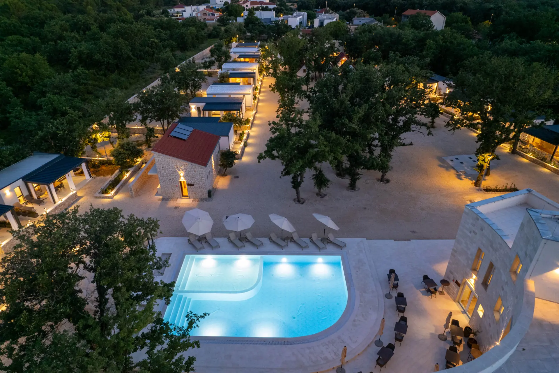 Aerial view of a swimming pool in a campground at night surrounded by trees and buildings. The swimming pool is clear blue and the buildings are mostly dark. The trees are green and some of them have lights on. This image is from Dionis Camping Zaton, a campground in Zaton, Croatia.