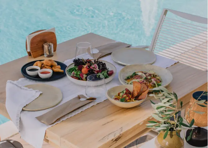 A wooden table with plates of food on it next to a swimming pool. The table is set for a meal with plates of pasta, salad, and bread. There are also drinks in glasses on the table. The swimming pool is blue and there are palm trees in the background.
                