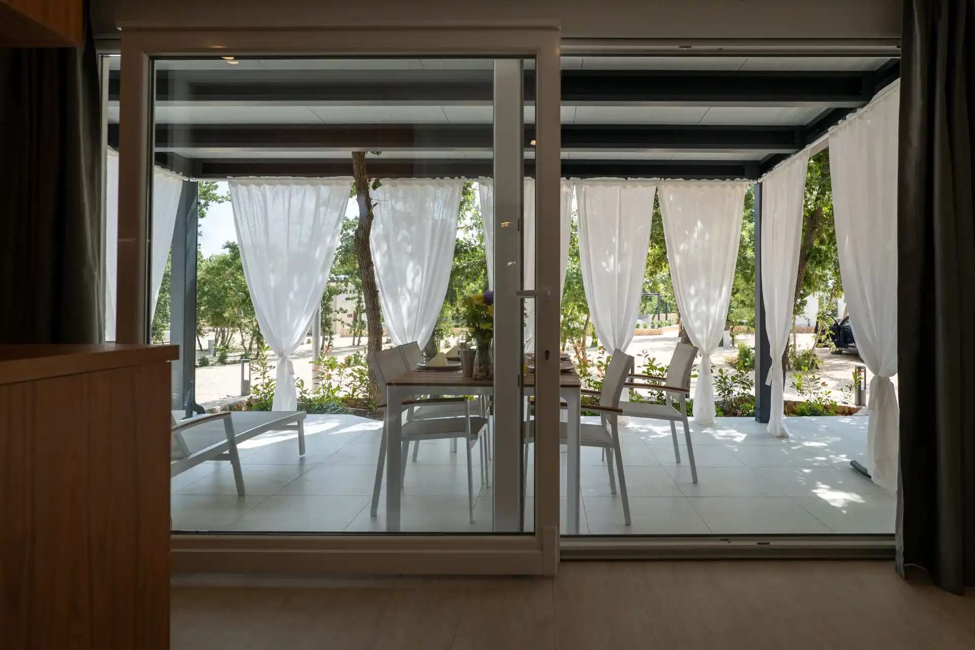 View from inside of mobile home to a terrace with white curtains and outdoor furniture