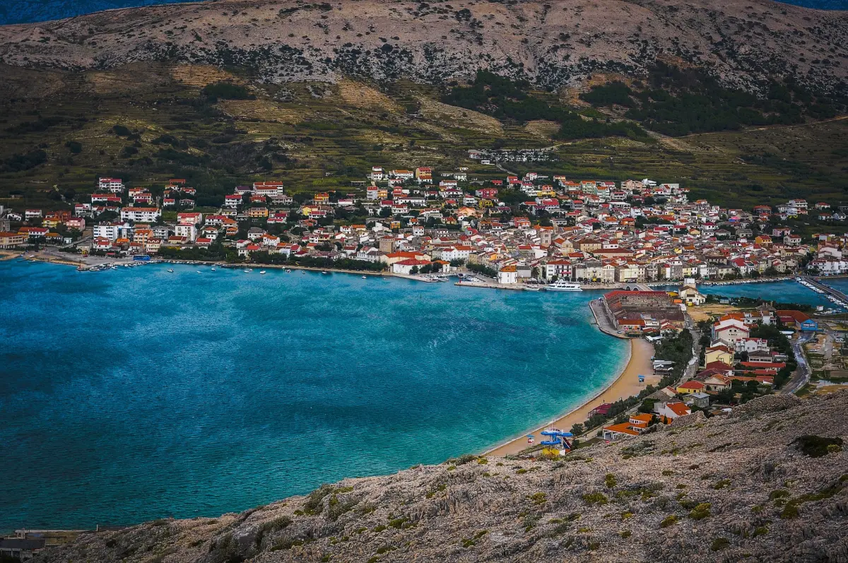 Mali grad Pag na obali Hrvatske. Grad je okružen vodom s tri strane, a u središtu grada nalazi se velika crkva. Kuće u gradu su većinom bijele, a ima i nešto drveća i grmlja.