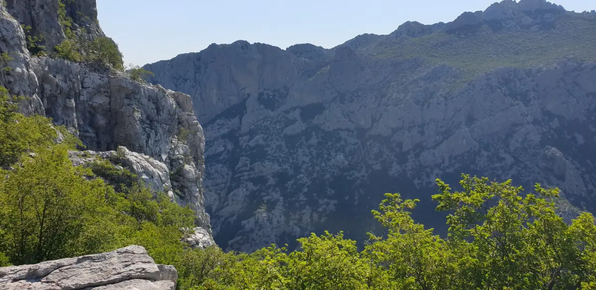 Blick auf eine Schlucht vom Gipfel eines Berges. Die Schlucht ist ein tiefer, schmaler Tal mit steilen Seiten. Die Wände der Schlucht haben verschiedene Brauntöne und Orangetöne, und auf ihnen wachsen Bäume und Büsche. Die Schlucht ist von Bergen umgeben, und im Hintergrund ist ein blauer Himmel zu sehen. Dieses Bild stammt aus dem Nationalpark Paklenica in Kroatien