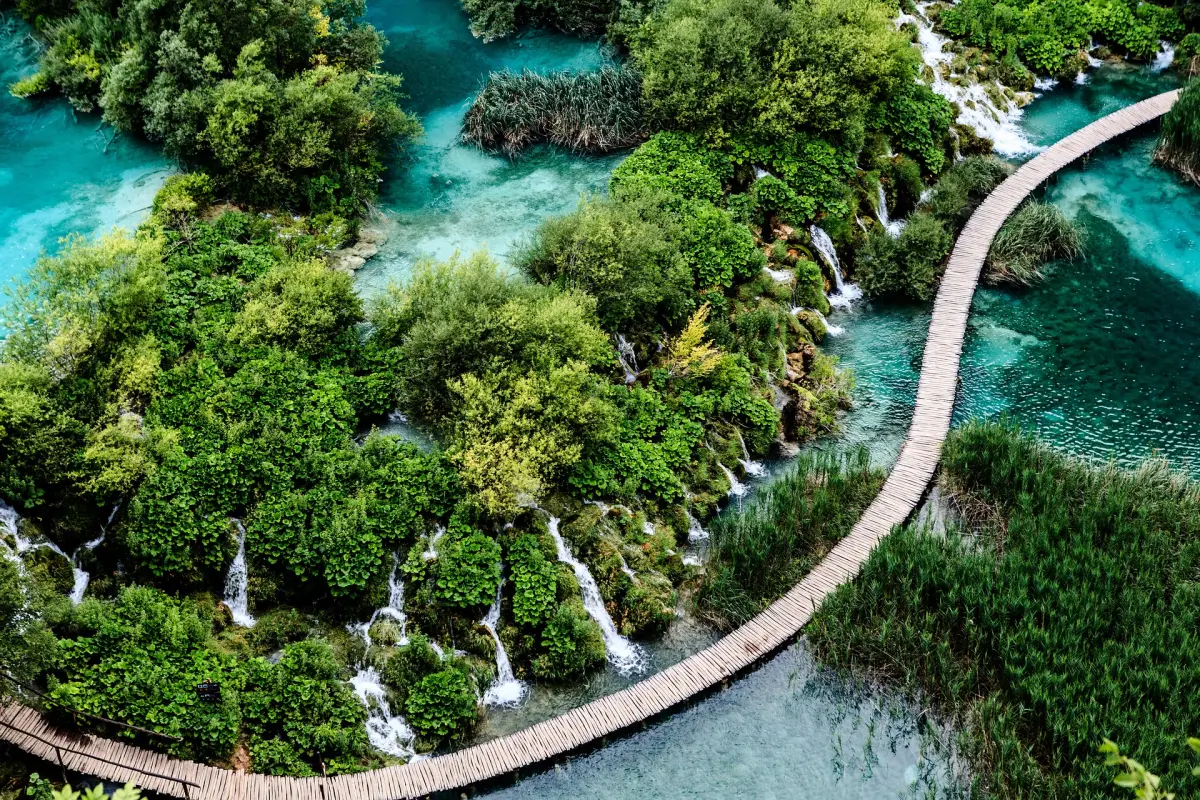 Ein hölzerner Steg über einen See im Nationalpark Plitvicer Seen in Kroatien. Der Steg ist von Bäumen und Bergen umgeben, und in der Ferne ist ein Wasserfall zu sehen