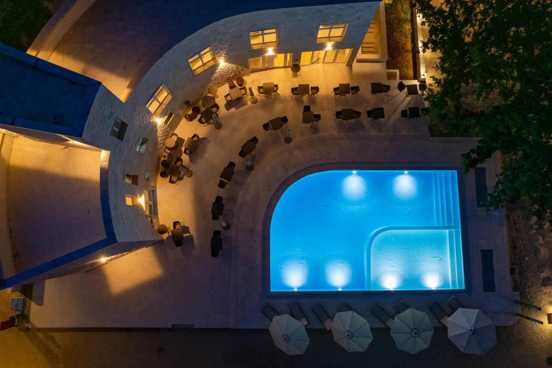 Aerial view of a swimming pool at night surrounded by chairs and umbrellas. The swimming pool is blue and there are chairs and umbrellas on the pool deck. There are lights on in the swimming pool and the surrounding area, making it a bright and inviting space